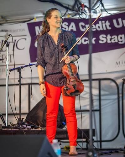 Rachel Schott standing on a festival stage, holding a violin and bow, smiling